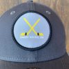 Grey patch on the front of a grey baseball hat featuring two crossed golden hockey sticks with writing below reading "Parry Sound, ON"