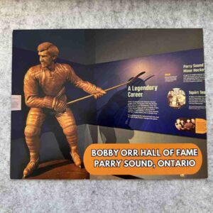 Photograph of a postcard featuring a bronze statue of Bobby Orr in front of exhibit display panels.