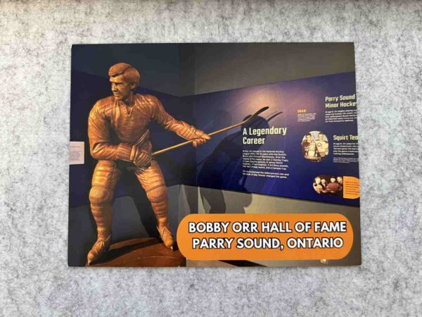 Photograph of a postcard featuring a bronze statue of Bobby Orr in front of exhibit display panels.